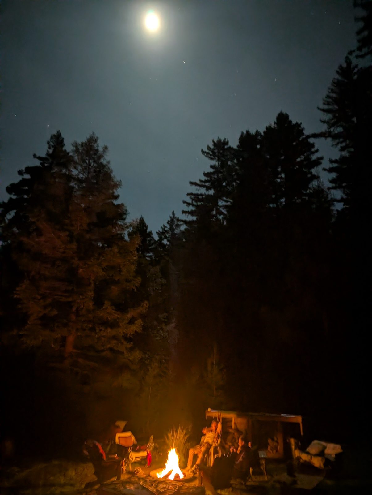 The moon over the treeline, a fire burning in the camp below
